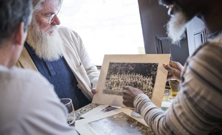 The image shows three individuals seated around a table, engaged in examining old photographs.